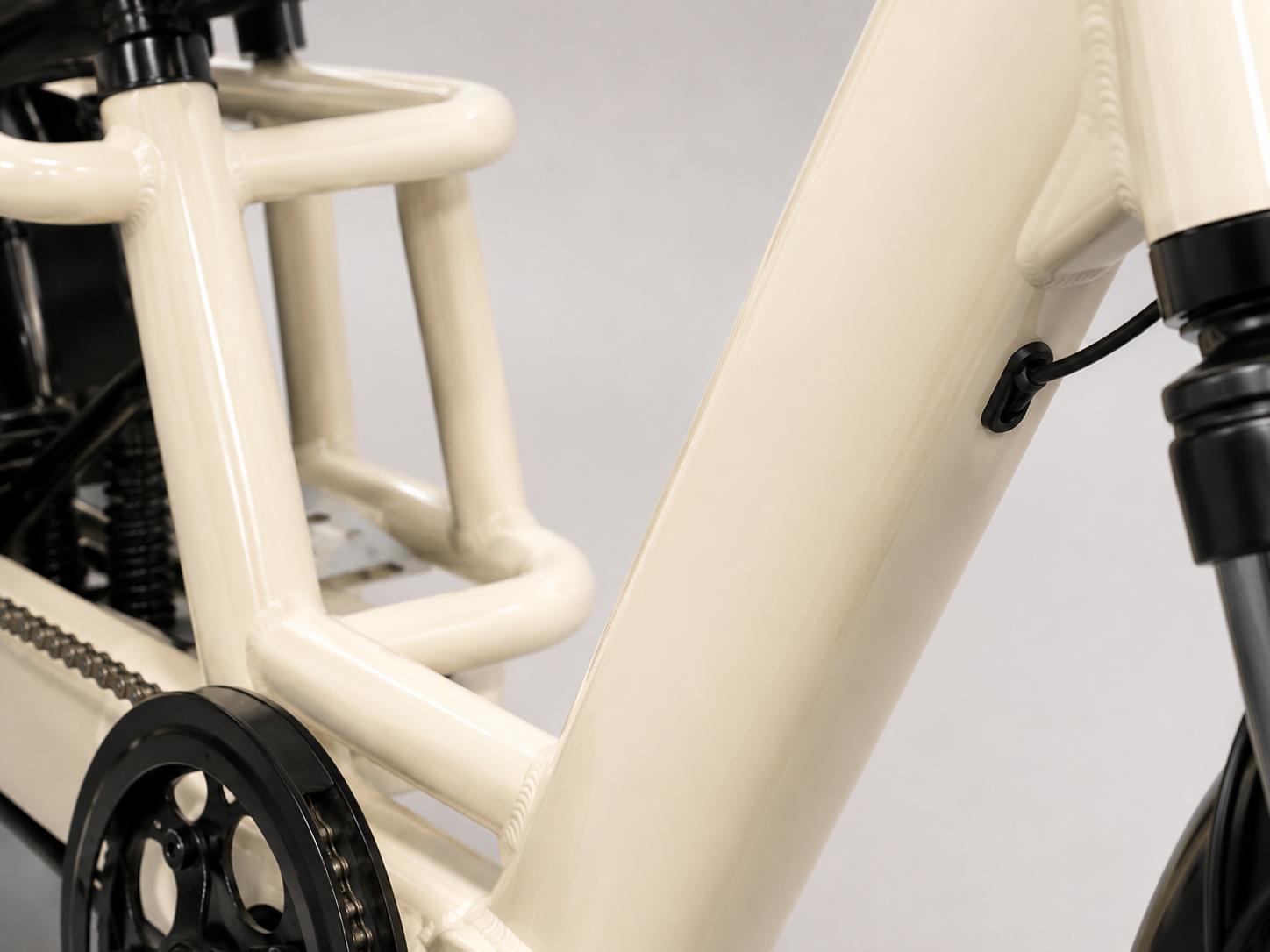 Close-up of a cream-colored electric cargo bike frame with black mechanical components, including a chainring, chain, and fork, set against a neutral light gray background.